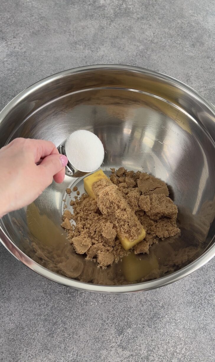 A hand holds a measuring spoon of white sugar over a metal bowl containing sticks of butter and piles of brown sugar, ready to be mixed. The bowl is set on a gray countertop.