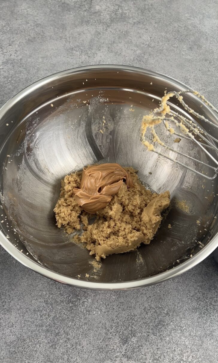 A metal mixing bowl contains cookie dough and a dollop of peanut butter, with a hand mixer resting on the edge. The bowl sits on a gray countertop.
