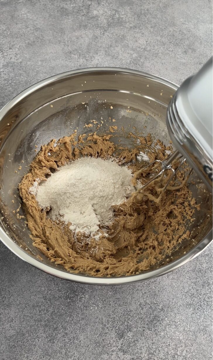 A metal mixing bowl with brown cookie dough and flour being mixed with a handheld electric mixer on a gray countertop.