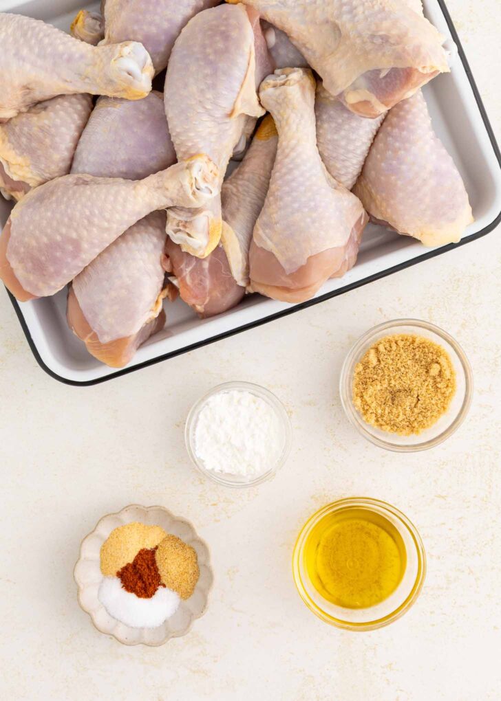 A tray of raw chicken drumsticks next to small bowls containing brown sugar, cornstarch, spices, and a bowl of olive oil on a white surface.