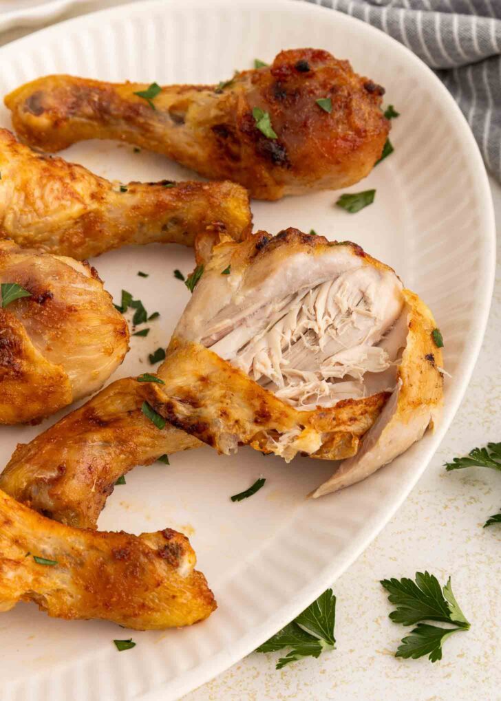 Four cooked chicken drumsticks on a white plate, garnished with chopped parsley. One drumstick is cut open, showing tender, juicy white meat inside. A few parsley leaves are scattered on the table nearby.