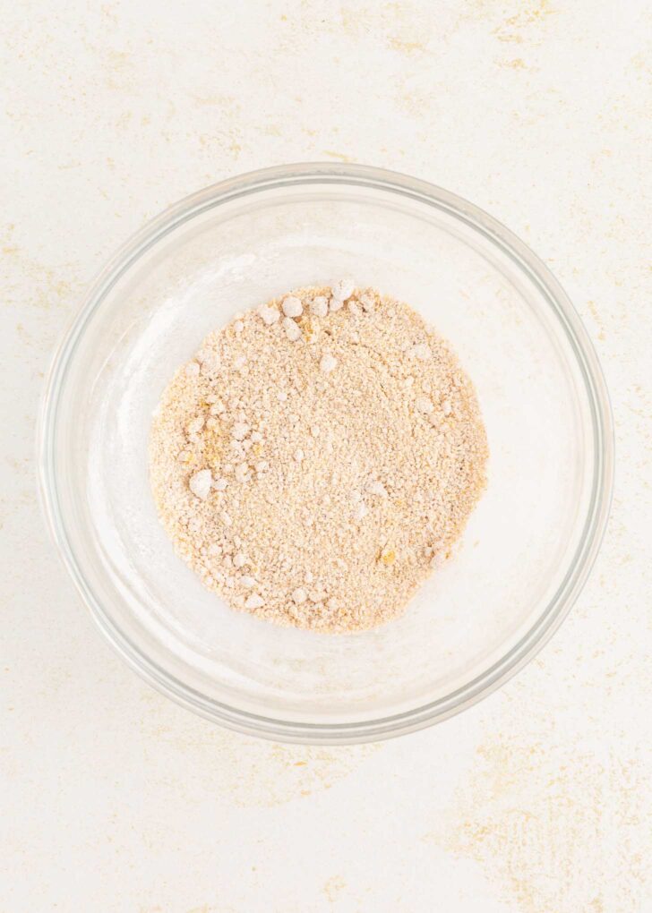 A clear glass bowl containing a light tan, crumbly dry mixture sits on a white surface.