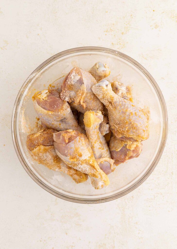 Raw chicken drumsticks coated in seasoning are arranged in a clear glass bowl on a light, textured surface.