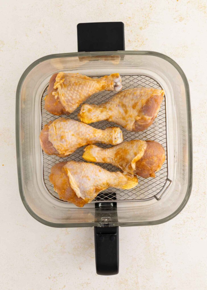 Six seasoned chicken drumsticks arranged in a single layer inside an air fryer basket, ready to be cooked. The basket has a metal mesh bottom and is viewed from above.