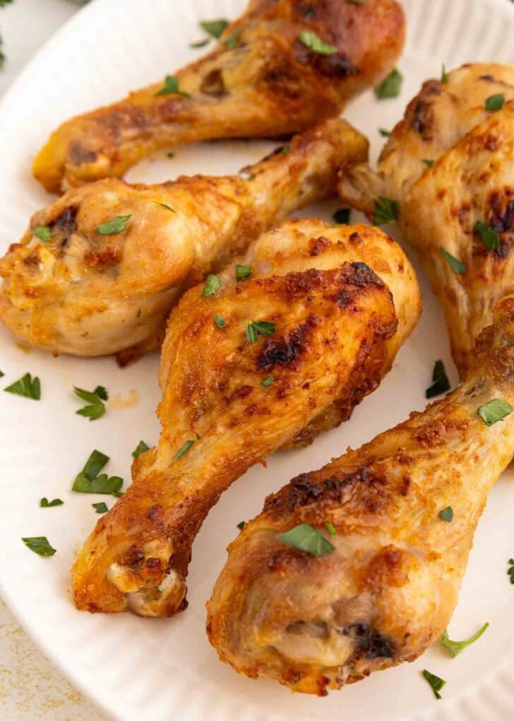 Five golden-brown chicken drumsticks garnished with chopped fresh herbs are arranged on a white plate. The chicken appears crispy and well-cooked.