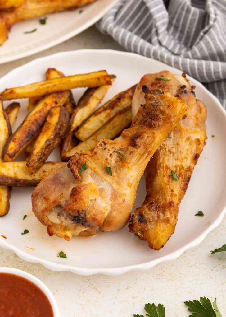 Two seasoned and chicken drumsticks served on a white plate with crispy potato wedges. A small bowl of sauce and fresh parsley leaves are visible nearby, with a gray-striped napkin in the background.