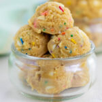 Scoops of edible cookie dough with colorful sprinkles are stacked in a small, clear glass bowl. Another bowl of cookie dough is blurred in the background.