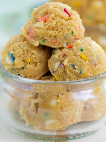 Scoops of edible cookie dough with colorful sprinkles are stacked in a small, clear glass bowl. Another bowl of cookie dough is blurred in the background.