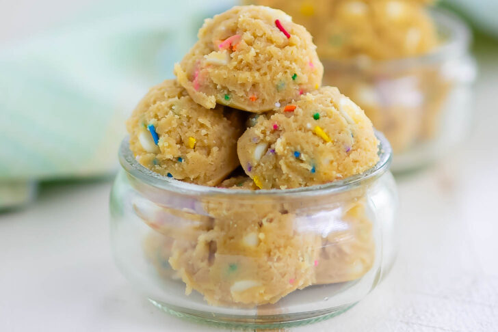 Scoops of edible cookie dough with colorful sprinkles are stacked in a small, clear glass bowl. Another bowl of cookie dough is blurred in the background.