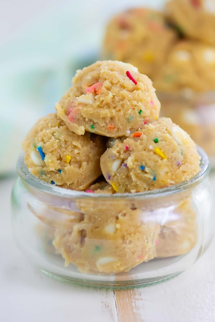 A small glass jar filled with scoops of cookie dough, featuring colorful sprinkles and white chocolate chips. More scoops are blurred in the background.