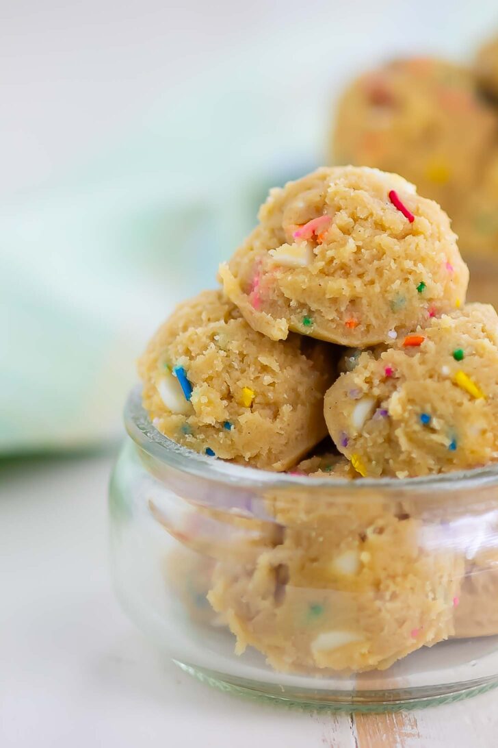 A small glass jar filled with scoops of cookie dough containing colorful sprinkles, with more dough pieces blurred in the background.