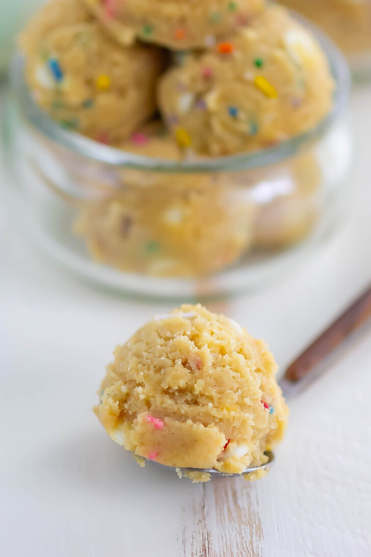A spoonful of cookie dough with colorful sprinkles sits in focus on a white surface, while more scoops of cookie dough are in a glass jar in the blurred background.