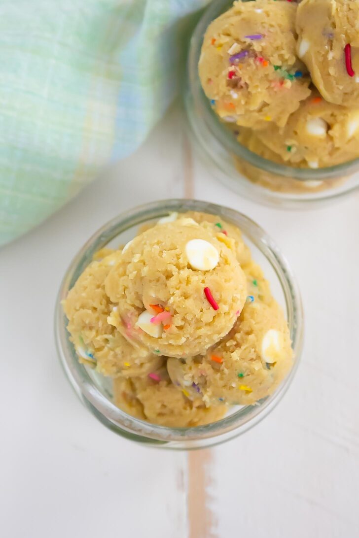 Overhead view of two glass jars filled with cookie dough balls containing white chocolate chips and colorful sprinkles, placed on a light surface next to a blue and green plaid cloth.