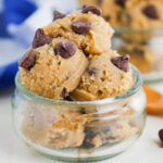 Scoops of chocolate chip cookie dough in a small glass jar, topped with chocolate chips. A wooden spoon and a blue and white cloth are in the background.