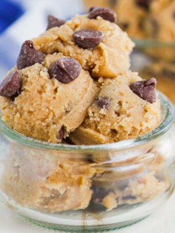 Scoops of cookie dough with chocolate chips in a small glass jar, with a wooden spoon and a blue-and-white cloth in the background.