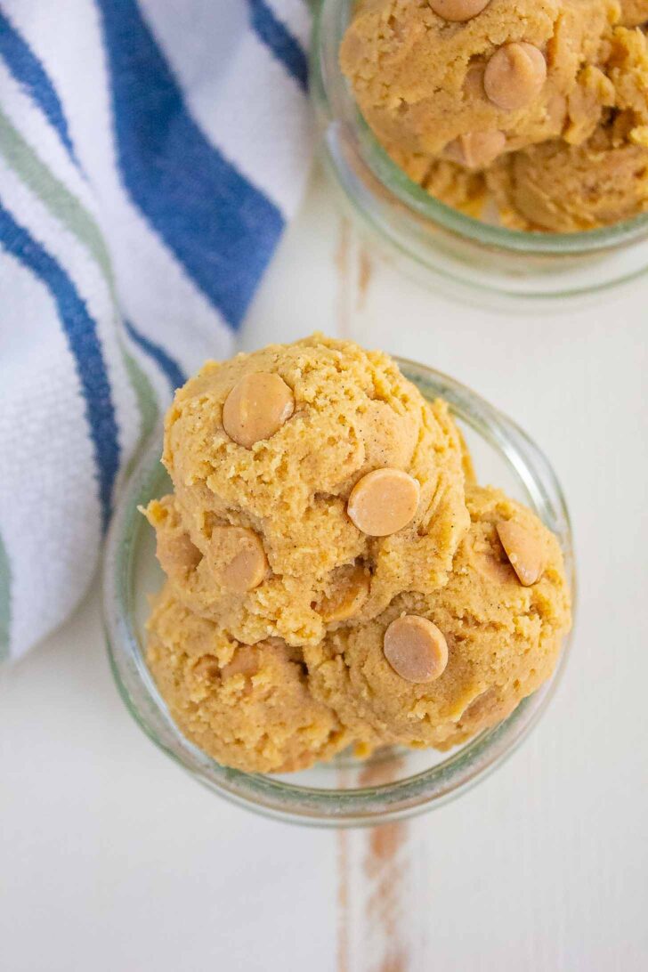Scoops of cookie dough with peanut butter chips in a small glass bowl, placed on a light surface next to a striped blue, green, and white cloth.