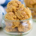 A small glass bowl filled with several scoops of cookie dough topped with peanut butter chips, with more cookie dough in the background and a striped cloth nearby.