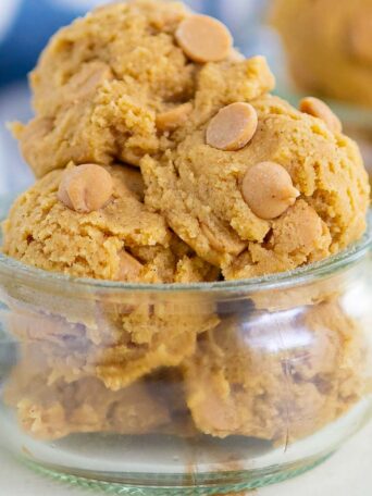 A small glass bowl filled with several scoops of cookie dough topped with peanut butter chips, with more cookie dough in the background and a striped cloth nearby.