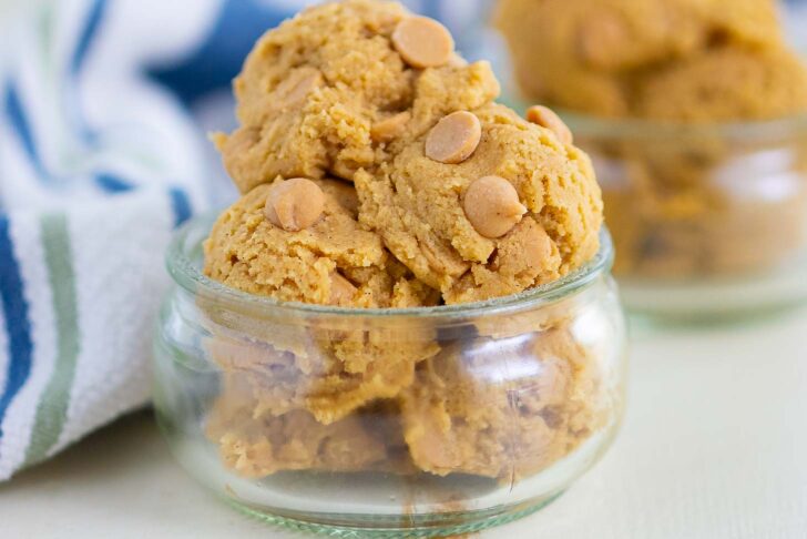 A small glass bowl filled with several scoops of cookie dough topped with peanut butter chips, with more cookie dough in the background and a striped cloth nearby.