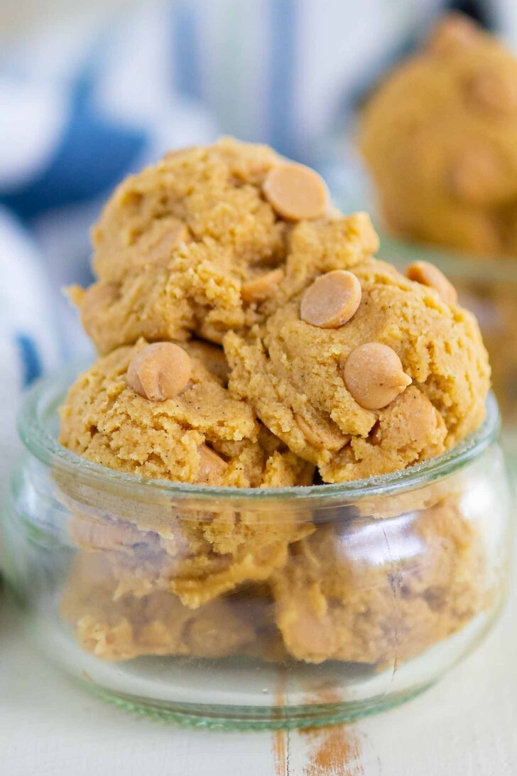 A small glass jar is filled with scoops of peanut butter cookie dough, each scoop studded with peanut butter chips. The jar sits on a light surface, and there are more scoops blurred in the background.