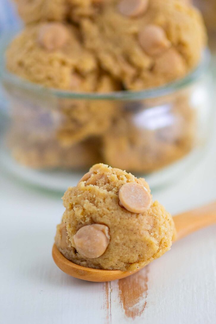A wooden spoon holds a scoop of cookie dough with peanut butter chips, with more dough and chips in a glass container in the background.