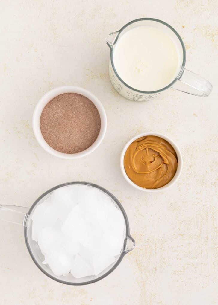 A top-down view of four containers: one with milk, one with hot chocolate mix, one with peanut butter, and one with ice cubes, all arranged on a light-colored surface.