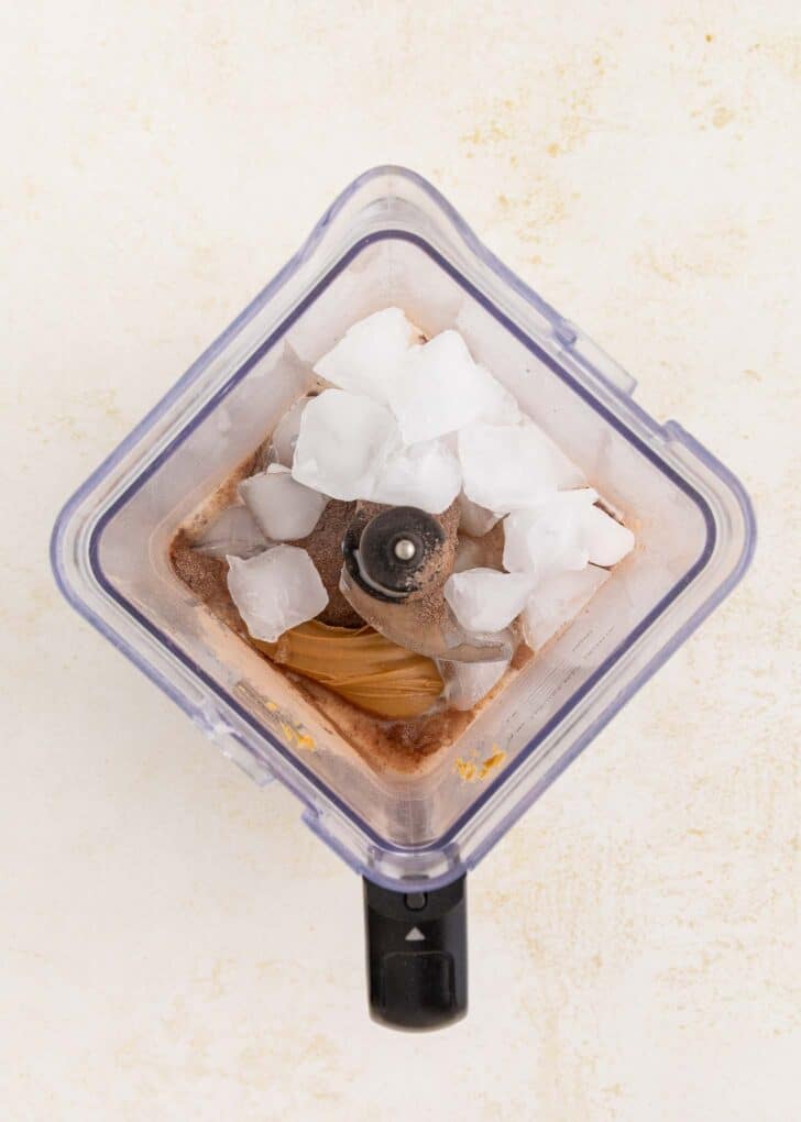 A top-down view of a blender containing ice cubes, hot chocolate mix, and peanut butter, ready to be blended.