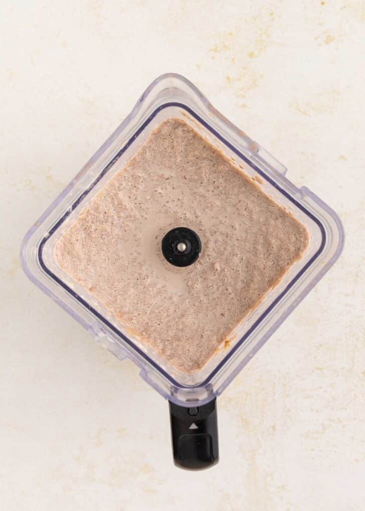 A top-down view of a blender filled with a smooth, light brown mixture, frozen hot chocolate, on a light-colored countertop.