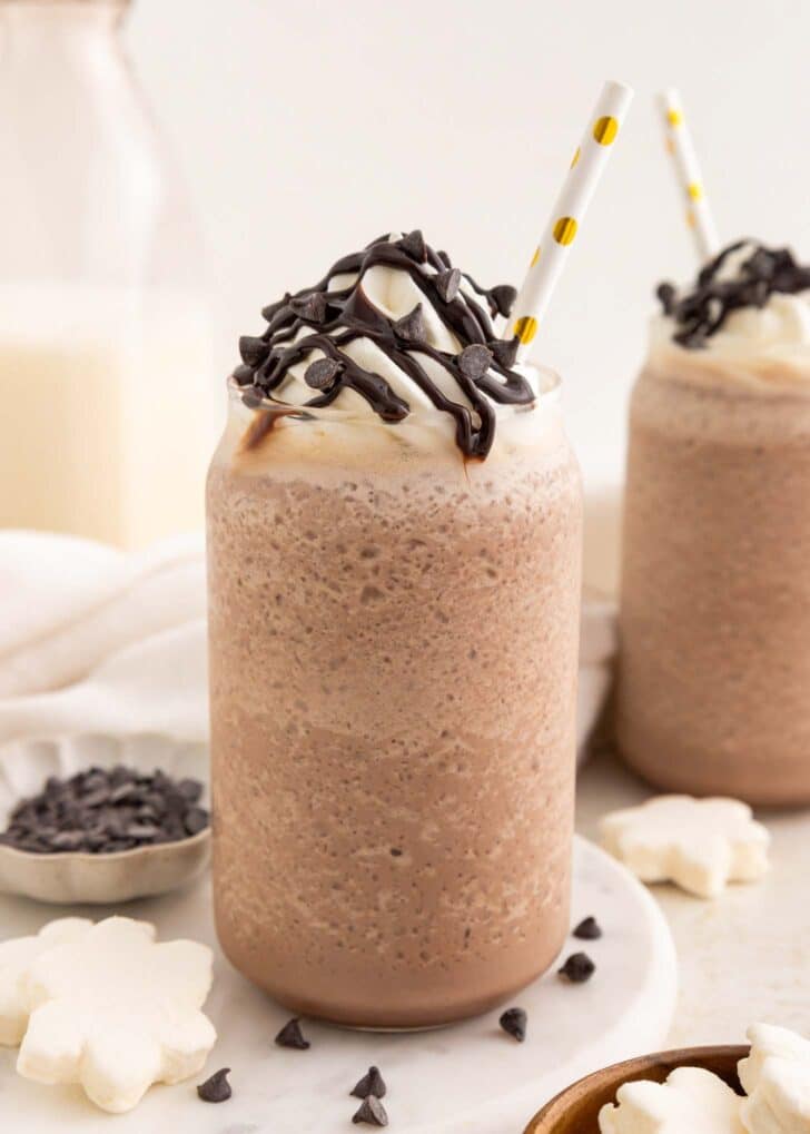 A glass of blended frozen hot chocolate topped with whipped cream, chocolate drizzle, and chocolate chips, with a yellow polka-dot straw. Another similar drink is in the background, along with milk, cookies, and chocolate chips.