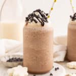Two glasses of blended frozen hot chocolate topped with whipped cream, chocolate drizzle, and chocolate chips. Each glass has a straw, and a milk bottle and small bowl of chocolate chips are in the background.