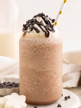 Two glasses of blended frozen hot chocolate topped with whipped cream, chocolate drizzle, and chocolate chips. Each glass has a straw, and a milk bottle and small bowl of chocolate chips are in the background.