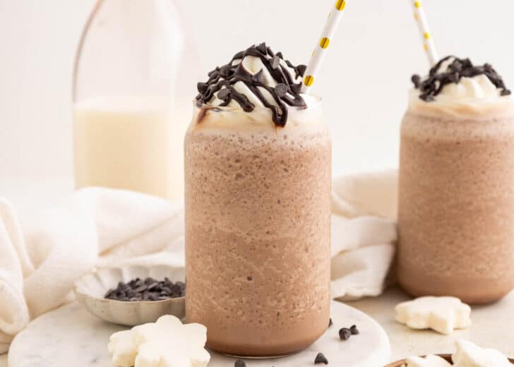 Two glasses of blended frozen hot chocolate topped with whipped cream, chocolate drizzle, and chocolate chips. Each glass has a straw, and a milk bottle and small bowl of chocolate chips are in the background.