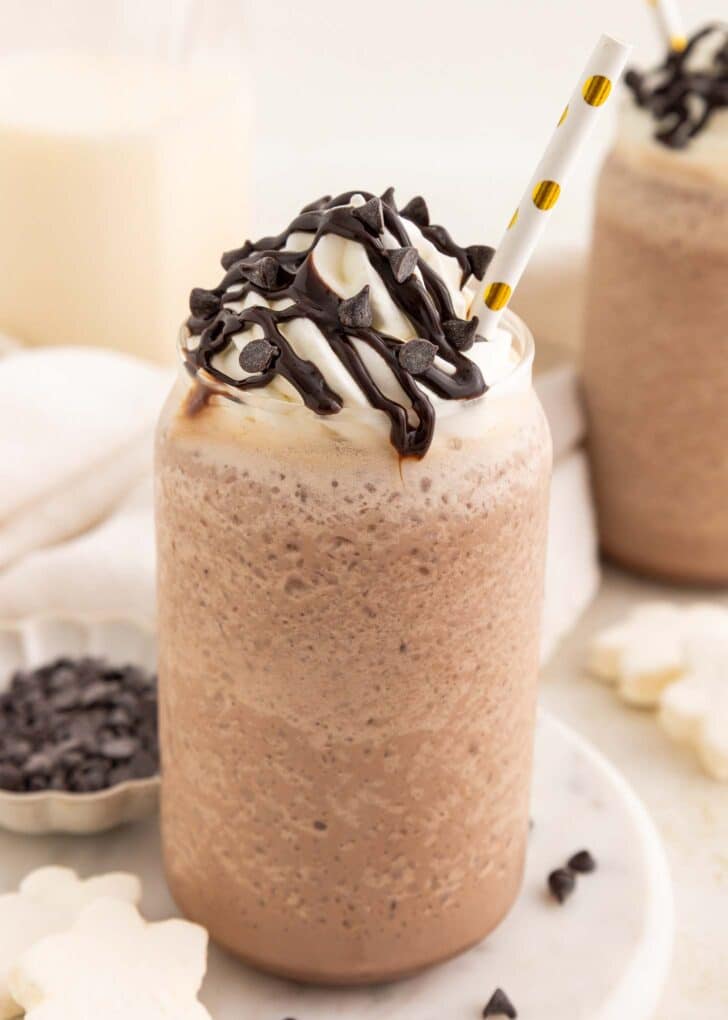 A glass of blended frozen hot chocolate topped with whipped cream, chocolate drizzle, and chocolate chips, with a white and gold straw. Another milkshake and chocolate chips are visible in the blurred background.