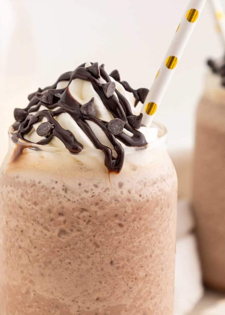 A close-up of a frozen hot chocolate in a glass, topped with whipped cream, chocolate syrup, and chocolate chips, with a white and yellow polka-dotted straw.