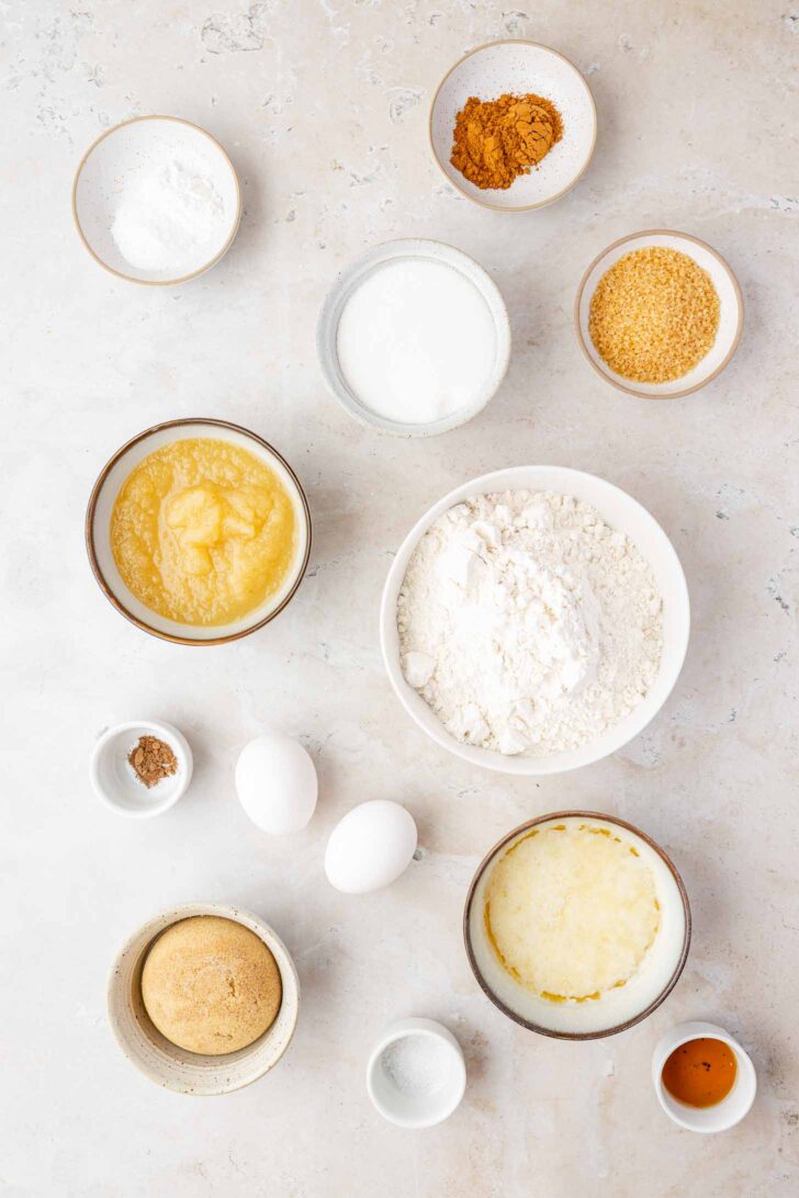Bowls on a countertop containing baking ingredients: flour, eggs, applesauce, brown sugar, granulated sugar, melted butter, cinnamon, ginger, milk, salt, and vanilla extract, all arranged neatly.
