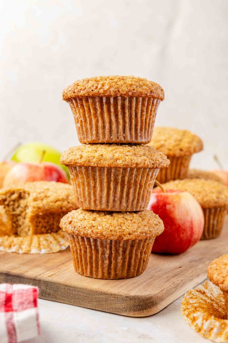 Three apple muffins are stacked on a wooden board, surrounded by red apples and more muffins, with one muffin partially unwrapped and a red-checked cloth visible in the corner.