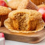 A close-up of an applesauce muffin with a bite taken out, resting on an unwrapped paper liner on a wooden board, surrounded by whole muffins and a red apple.