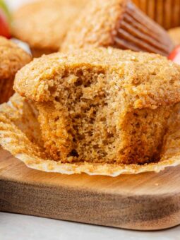 A close-up of an applesauce muffin with a bite taken out, resting on an unwrapped paper liner on a wooden board, surrounded by whole muffins and a red apple.