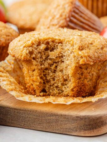 A close-up of an applesauce muffin with a bite taken out, resting on an unwrapped paper liner on a wooden board, surrounded by whole muffins and a red apple.