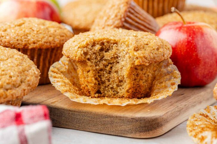 A close-up of an applesauce muffin with a bite taken out, resting on an unwrapped paper liner on a wooden board, surrounded by whole muffins and a red apple.