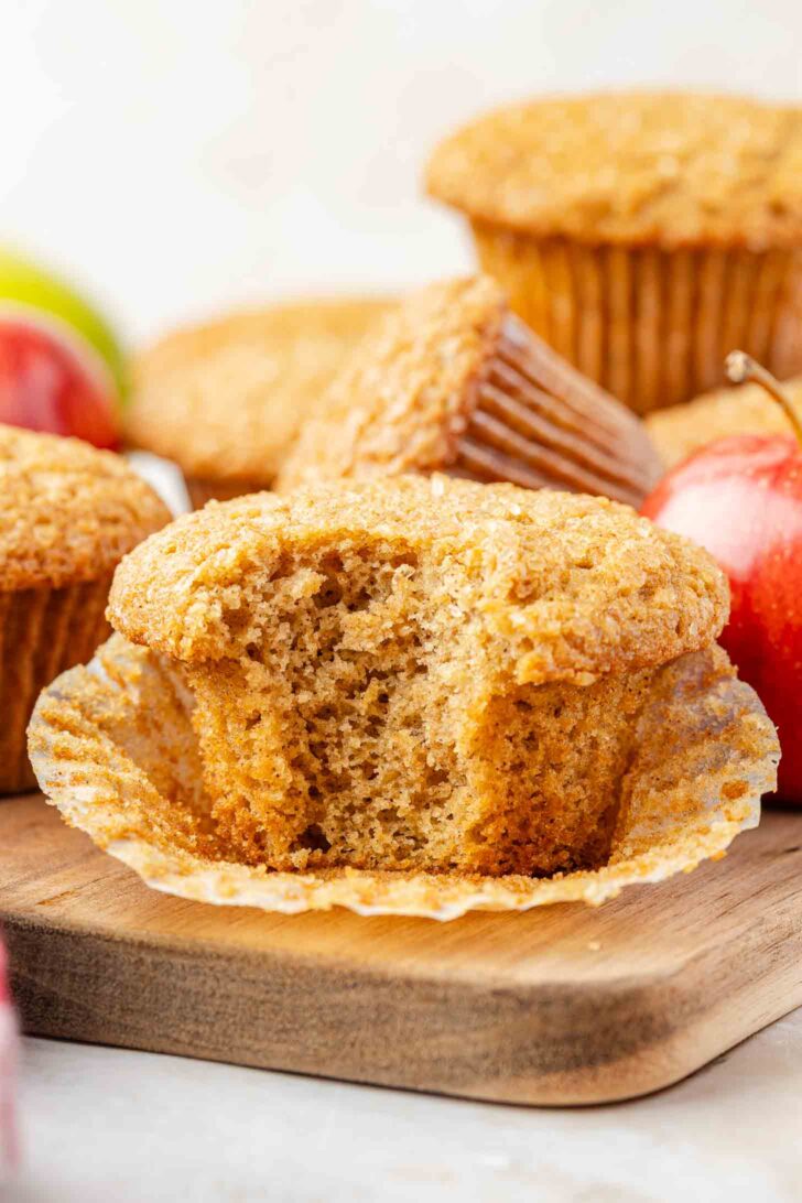A close-up of a partially eaten muffin with a golden brown top, resting in an open paper liner on a wooden surface. Whole muffins and red apples are blurred in the background.