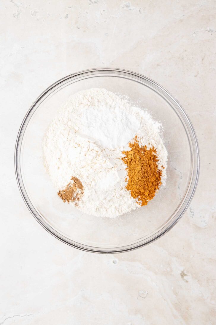 A clear glass bowl containing white flour and two small piles of brown and tan spices on a light-colored countertop.