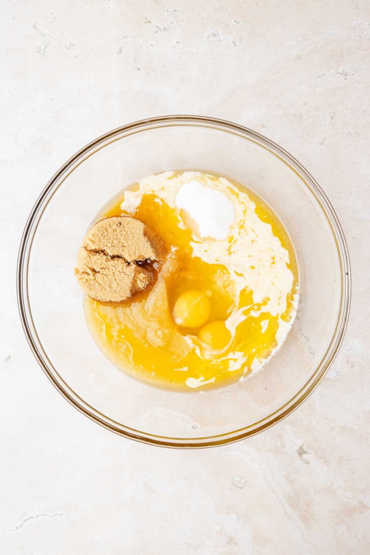 A glass mixing bowl containing brown sugar, two eggs, melted butter, and flour on a light-colored countertop, viewed from above.