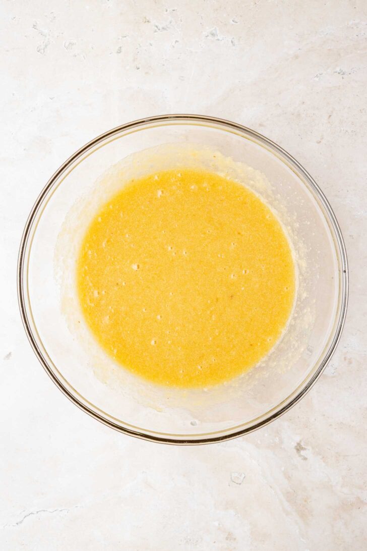 A clear glass bowl containing a smooth, yellow batter sits on a light-colored, textured surface.