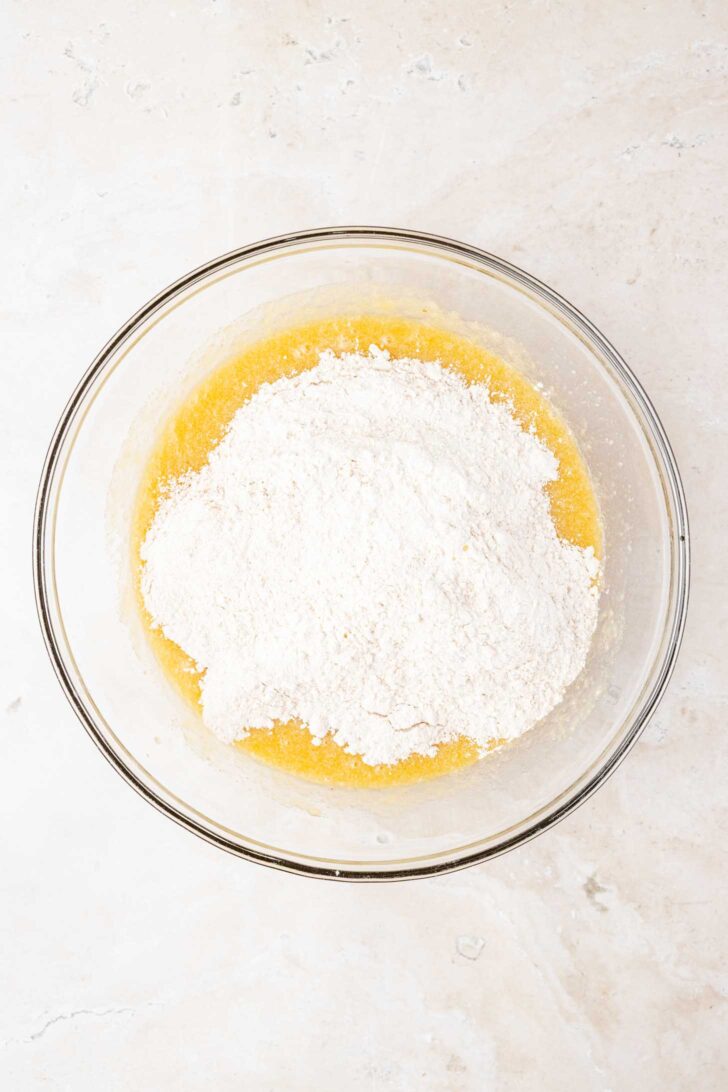 A glass mixing bowl with flour sitting on top of a yellow liquid mixture, placed on a light-colored marble surface.