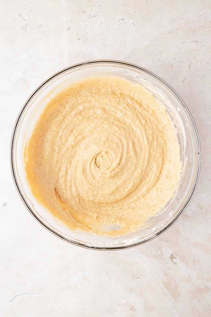 A glass mixing bowl filled with smooth, light beige cake batter sits on a light-colored marble surface. The batter has been mixed into a creamy, swirled texture.