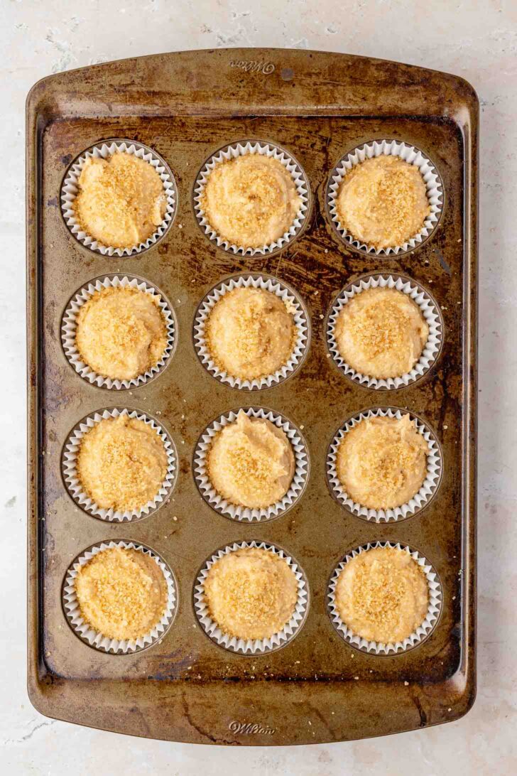 A metal muffin tin holds twelve cupcake liners filled with unbaked batter, each topped with a crumbly streusel, ready to be baked. The tin rests on a light-colored surface.