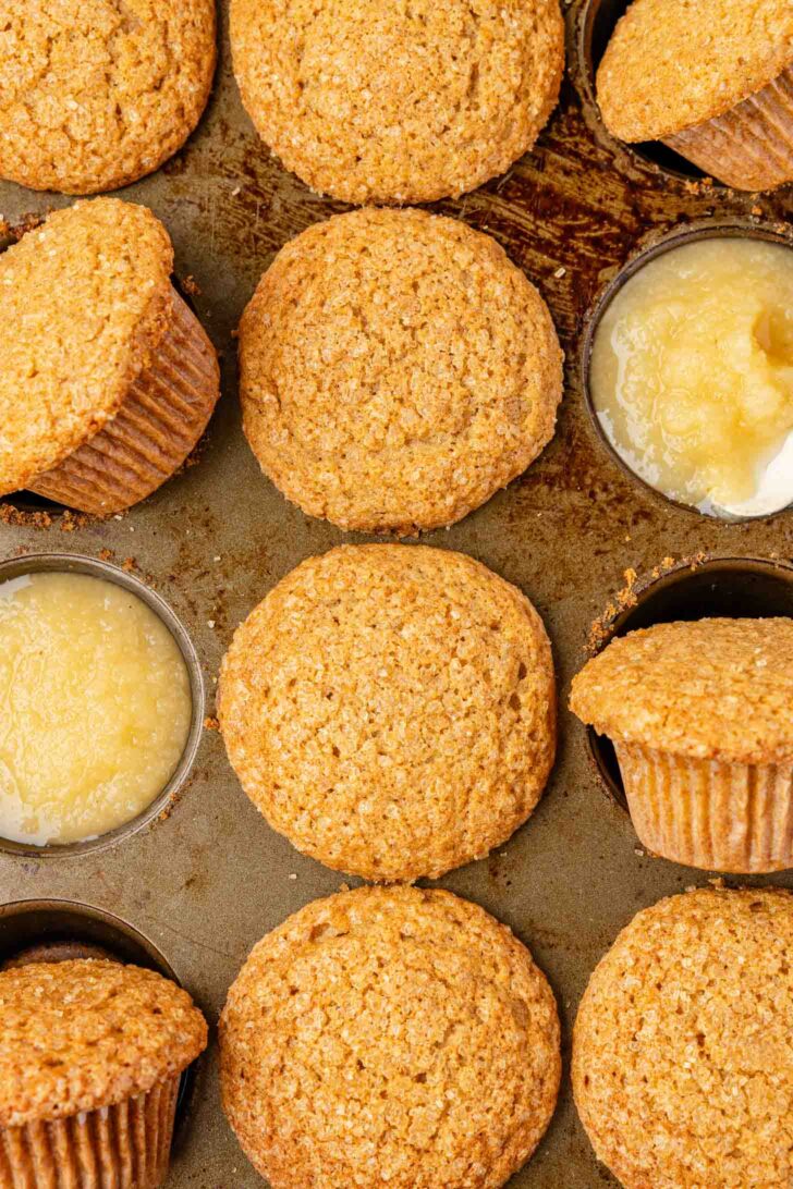 A close-up of several golden-brown muffins in a muffin tin, with two cups filled with applesauce instead of muffins. Some muffins are slightly tilted or out of their cups.