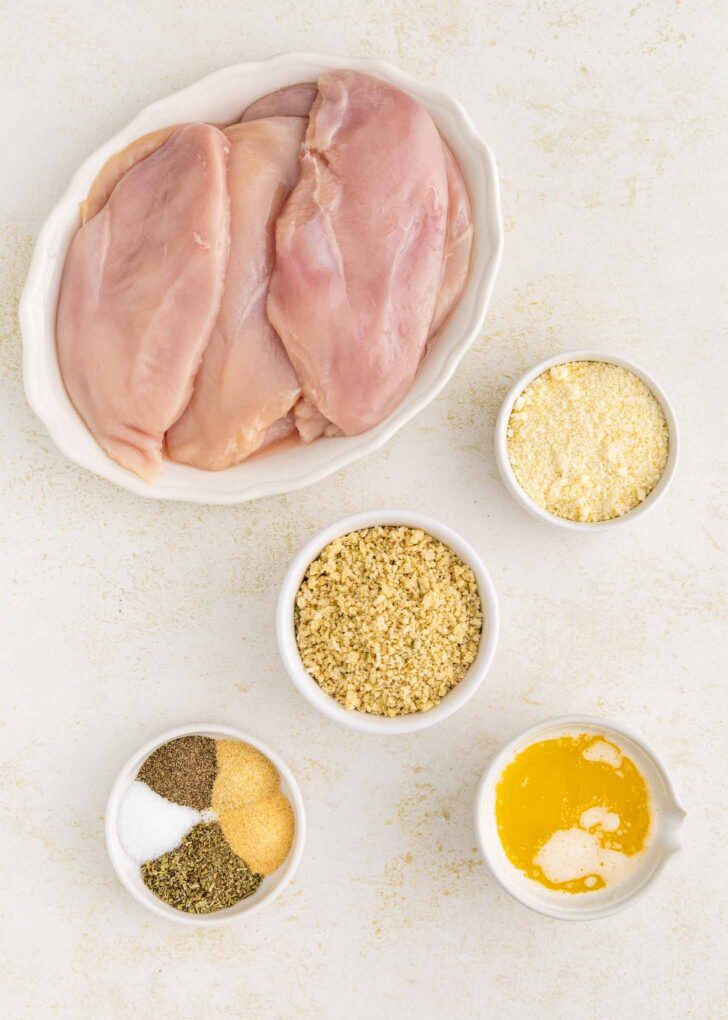 Raw chicken breasts in a white dish, surrounded by small bowls containing breadcrumbs, grated Parmesan cheese, melted butter, and assorted spices on a light surface.