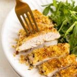 A fork picking up a slice of golden, breadcrumb-coated chicken breast next to a serving of fresh arugula on a white plate.